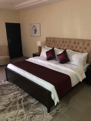 A bedroom with a large bed, a headboard, a bedside table with a lamp, a chair, and a door.