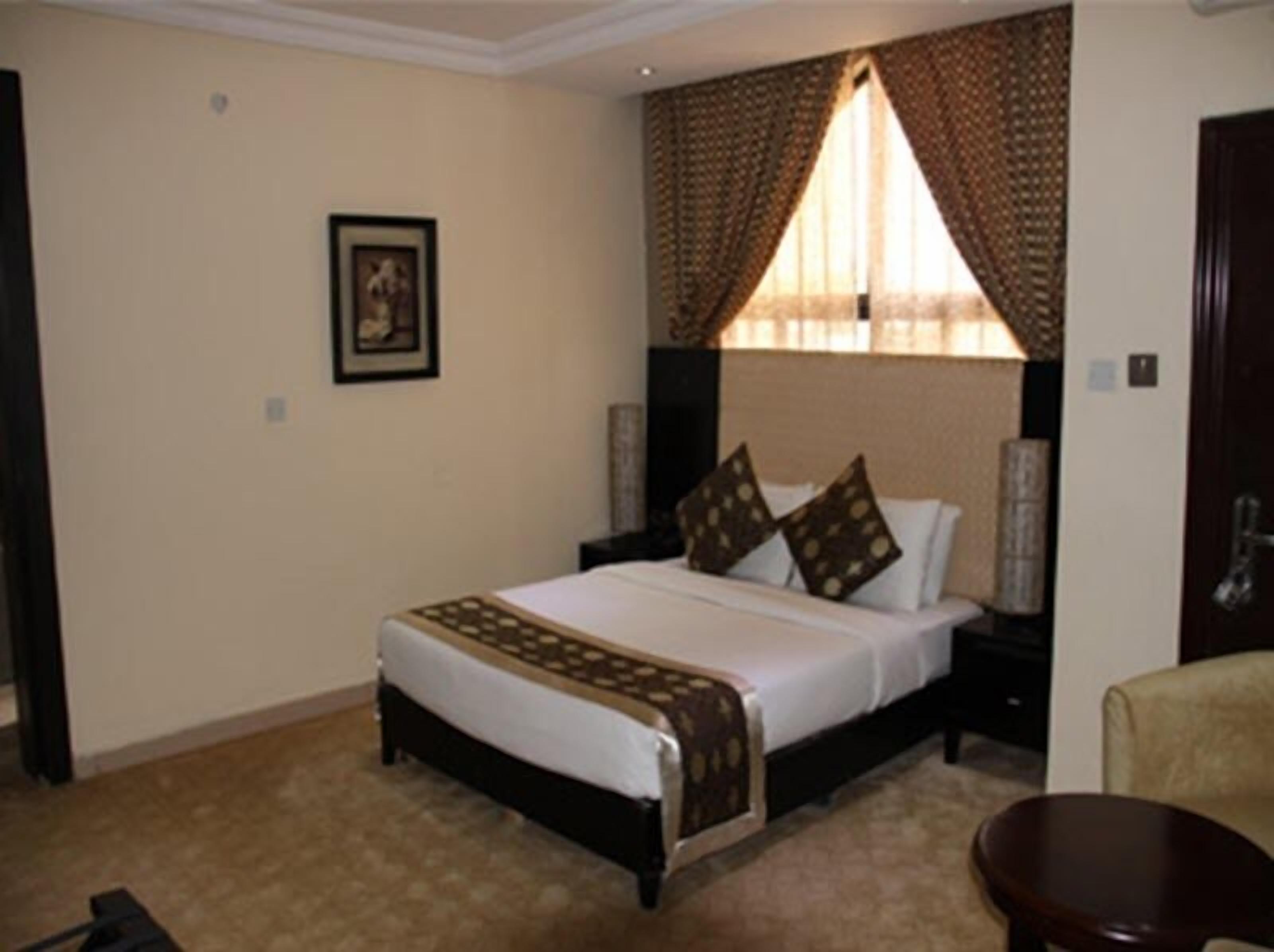 A hotel room with a large bed, a headboard with curtains, a framed picture on the wall, and a small table.
