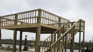 Eine Holzkonstruktion mit Treppe und Geländern, vermutlich eine Aussichtsplattform oder ein Pavillon, gelegen auf sandigem Untergrund mit einem Gewässer im Hintergrund.
