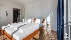 Un dormitorio con cabecera de cama de madera, ropa de cama blanca, una mesita de noche con lámpara y un pequeño objeto decorativo.