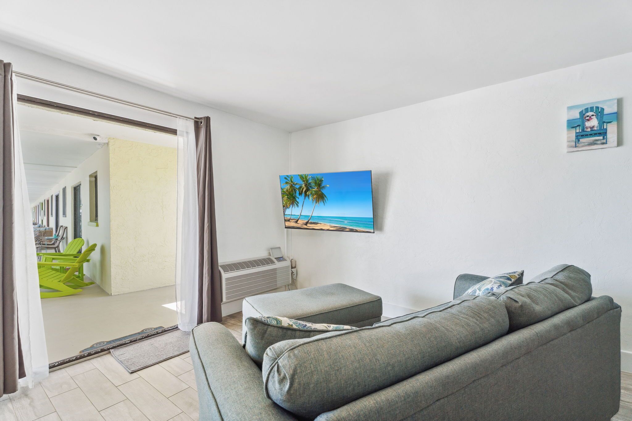 A living room with a grey sofa, a flat-screen TV on the wall, and a view of a balcony with green chairs.