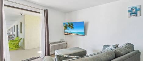 A living room with a grey sofa, a flat-screen TV on the wall, and a view of a balcony with green chairs.
