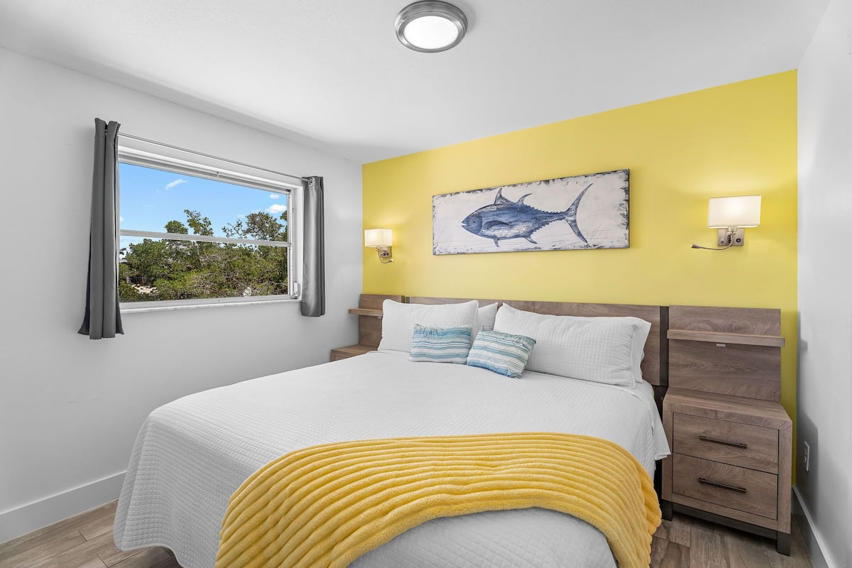 A bedroom with a bed, a nightstand, a window with curtains, a wall-mounted artwork of a fish, and a yellow throw blanket.