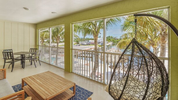 Een balkon met een houten tafel en stoelen, een hangstoel en uitzicht op palmbomen en een zwembad.