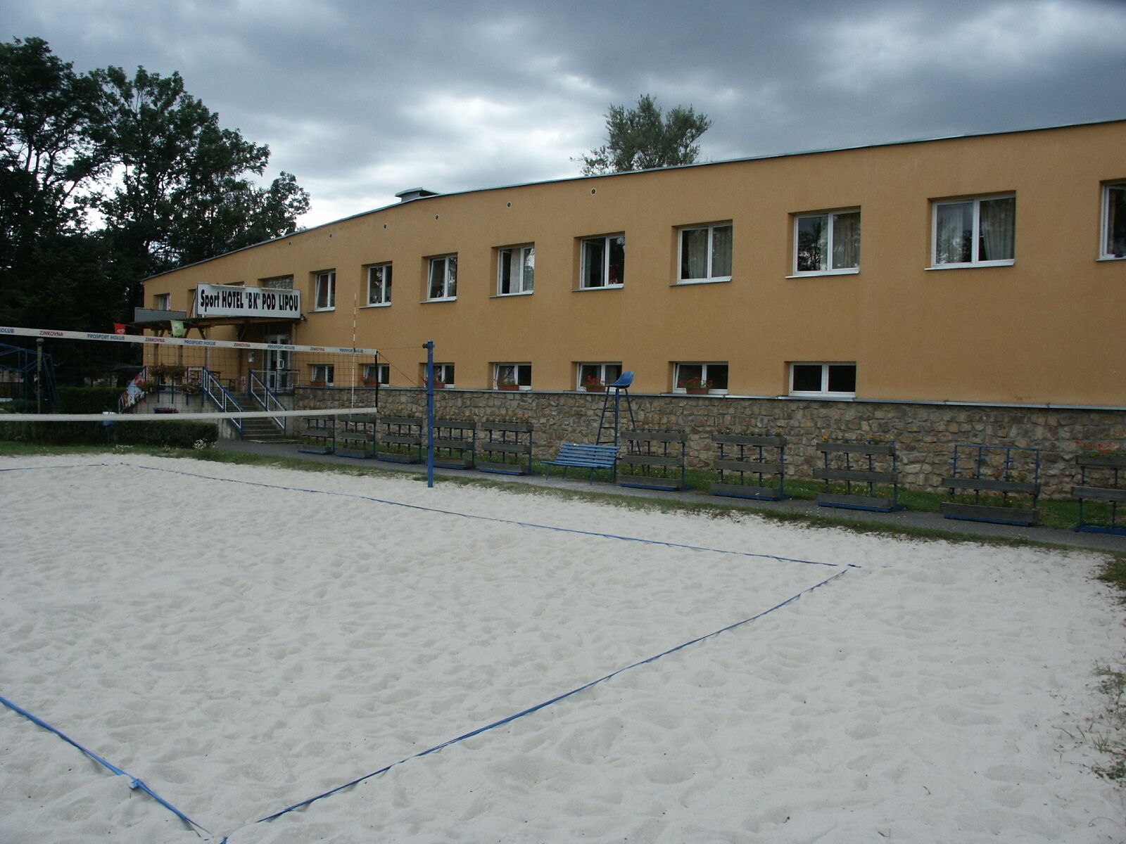 A sandy beach volleyball court adjacent to a building with multiple windows and a sign reading "Sport Hotel 'BK PODIPOU'.