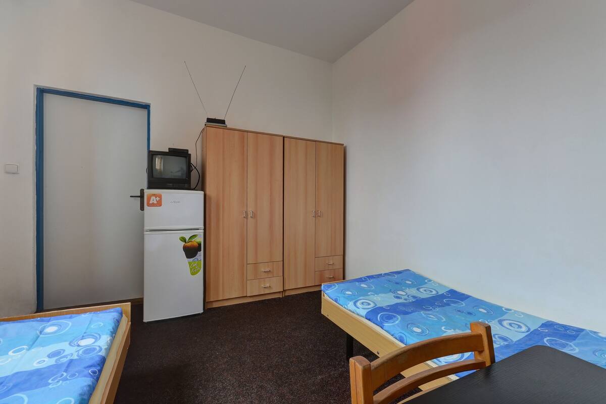 A small, compact room with two single beds, a wooden wardrobe, a small refrigerator, and a television on top of the wardrobe.