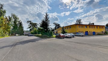 Un estacionamiento con varios coches, un edificio amarillo con puertas azules y árboles al fondo.