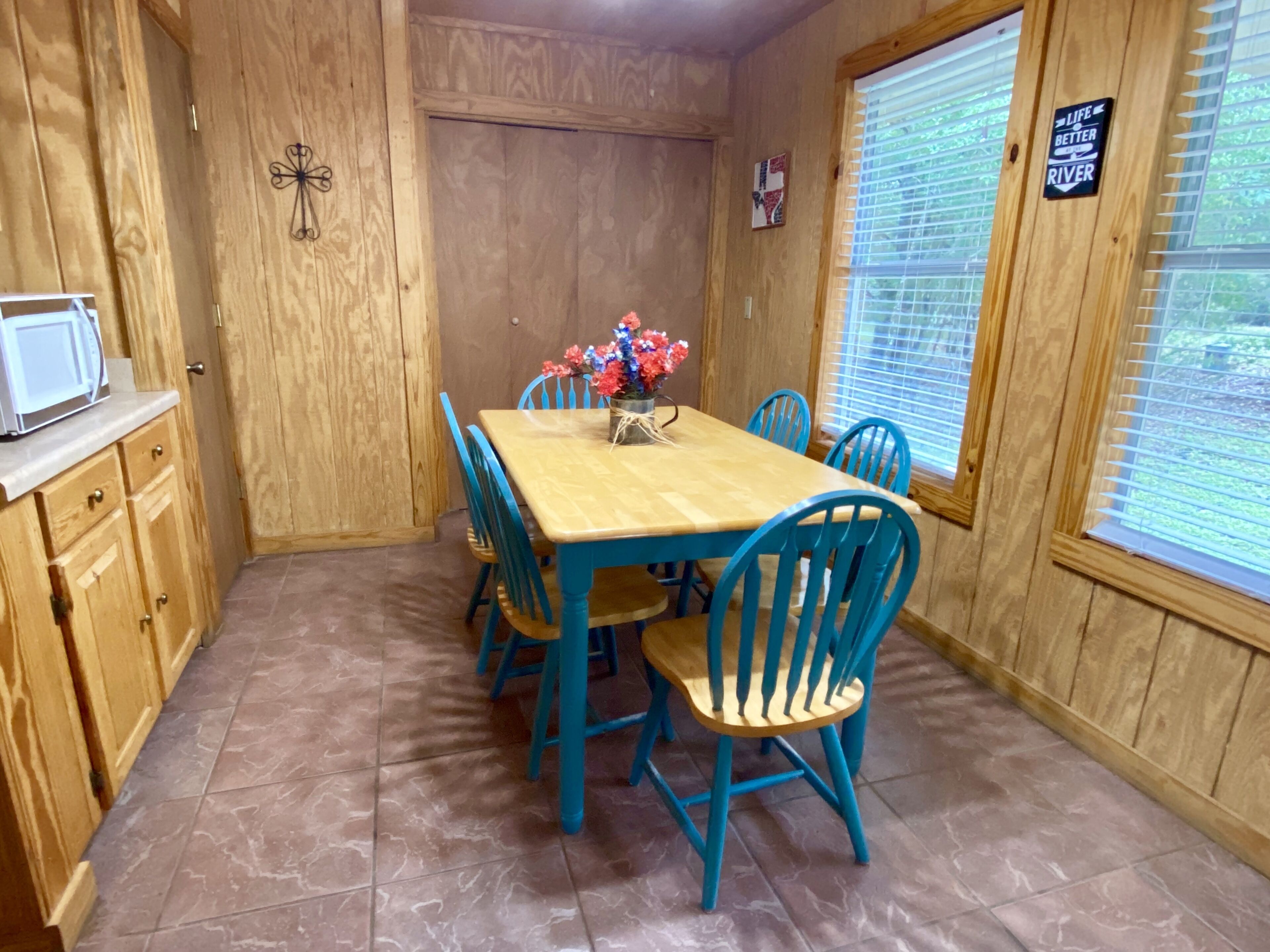 Una habitación con paneles de madera, una mesa de comedor, sillas azules, un jarrón con flores, un microondas y un cartel en la ventana.