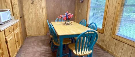 Una habitación con paneles de madera, una mesa de comedor, sillas azules, un jarrón con flores, un microondas y un cartel en la ventana.