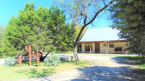 A rustic building with a stone facade and a metal roof, surrounded by trees and a gravel path. - Float 2 Cabin ~ Private Frio River Access @ Comanche Crossing, Cowboy Style! (Concan)