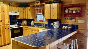 A kitchen with wooden cabinets, a blue-tiled countertop, a stove, a sink, and a window with curtains. - Float 2 Cabin ~ Private Frio River Access @ Comanche Crossing, Cowboy Style! (Concan)