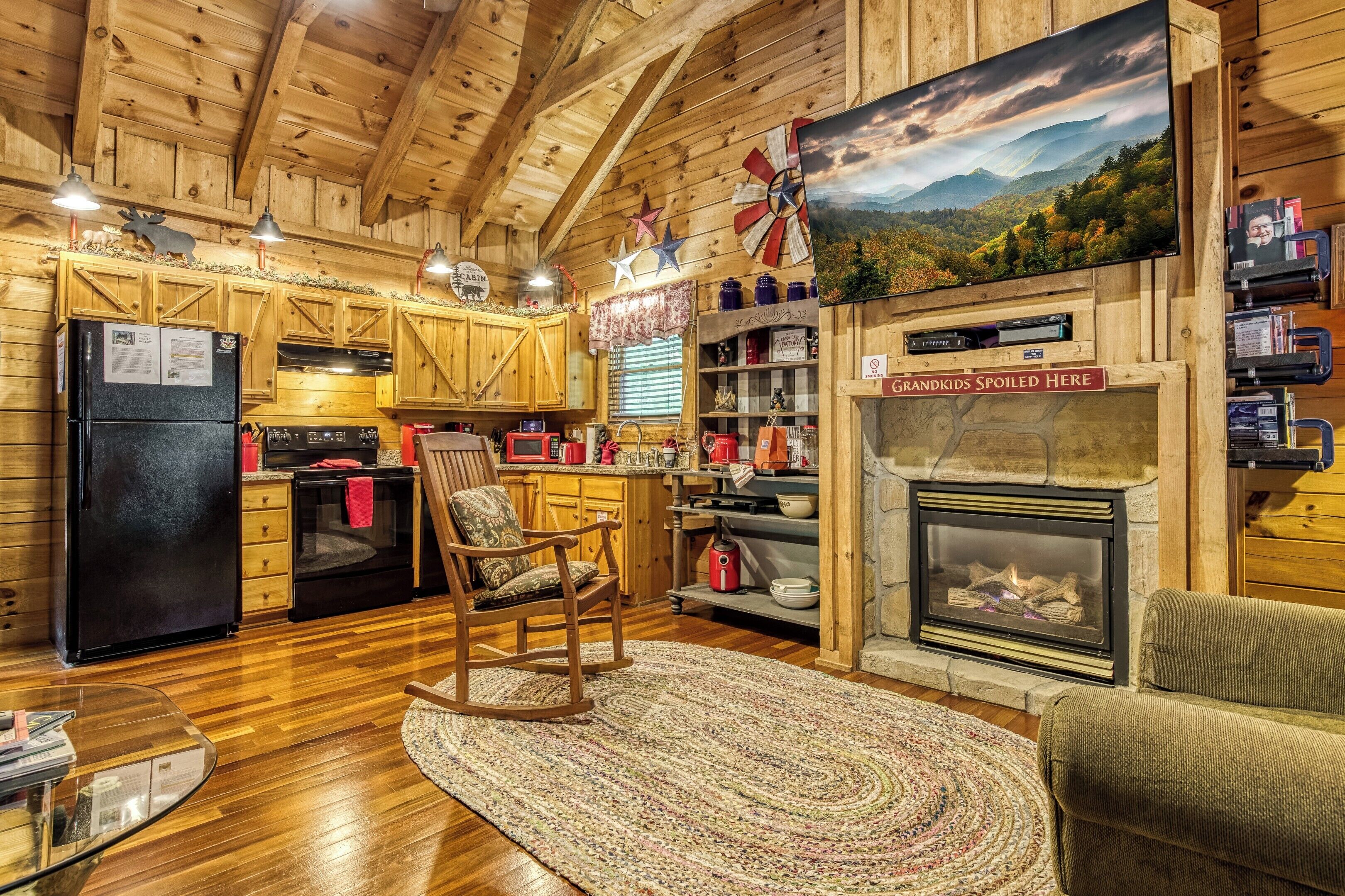 A cozy cabin interior with wooden walls, a fireplace, a kitchen with wooden cabinets, and a rocking chair.