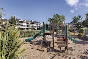 A playground with a slide and climbing structures, surrounded by a gravel area and greenery, with a building in the background.