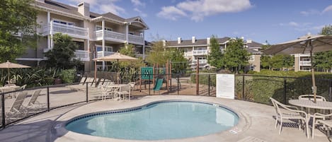 A swimming pool with lounge chairs and umbrellas, surrounded by a fence and a playground.