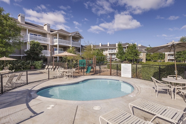 A swimming pool with lounge chairs and umbrellas, surrounded by a fence and a playground. - Beautiful Resort in CA Wine region - Windsor! (Windsor)