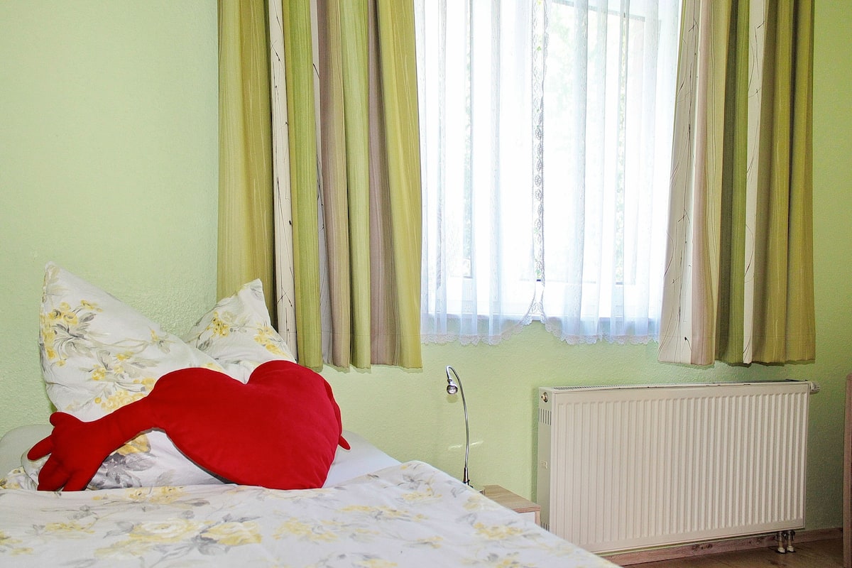 Une chambre à coucher avec un lit, une literie à motifs floraux, un coussin rouge en forme de cœur, un radiateur et des rideaux verts.