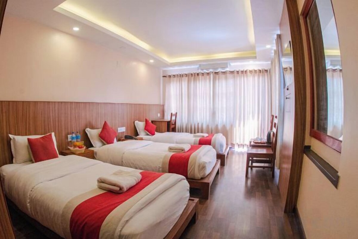 A hotel room with three beds, a wooden headboard, a window with curtains, and a small table with a lamp.