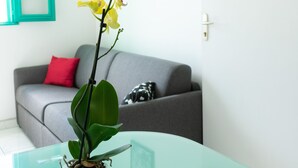 A modern living room with a glass table, a potted orchid, a grey sofa with a red pillow, and a teal-framed window.