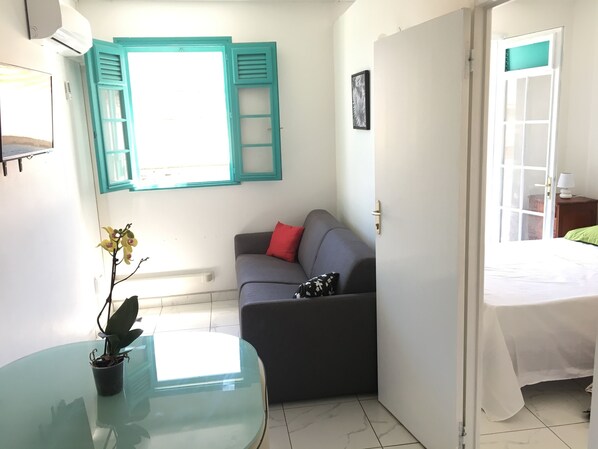 Habitación compacta con una mesa de vidrio, un sofá gris con un cojín rojo, una planta en maceta y una ventana con contraventanas color turquesa.