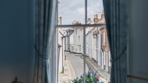 Une vue à travers une fenêtre sur une rue étroite bordée de bâtiments blancs, avec un lampadaire et une jardinière.