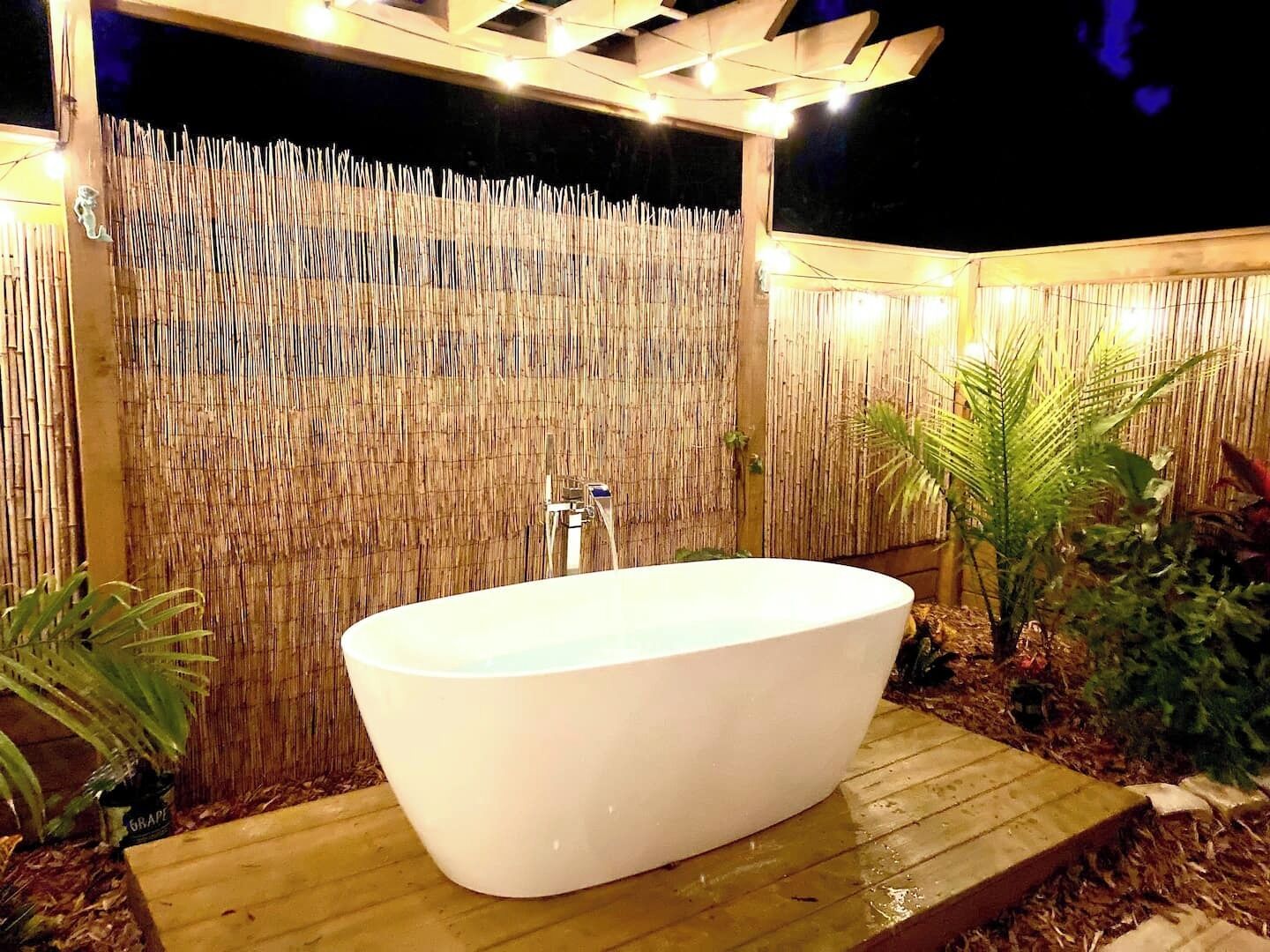 A freestanding bathtub in an outdoor setting with a bamboo backdrop and string lights.