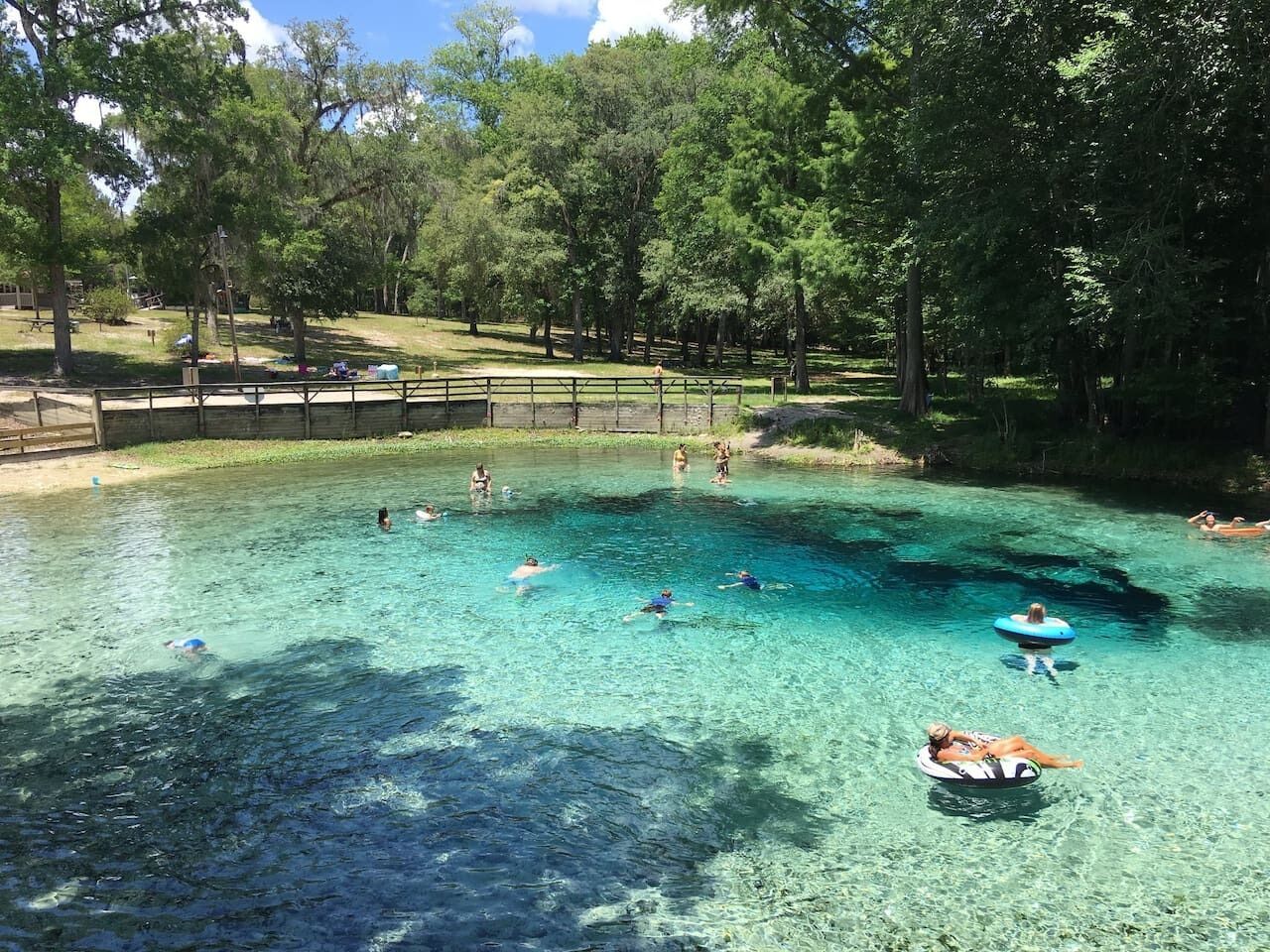 A clear, shallow water body surrounded by trees, with people swimming and floating.
