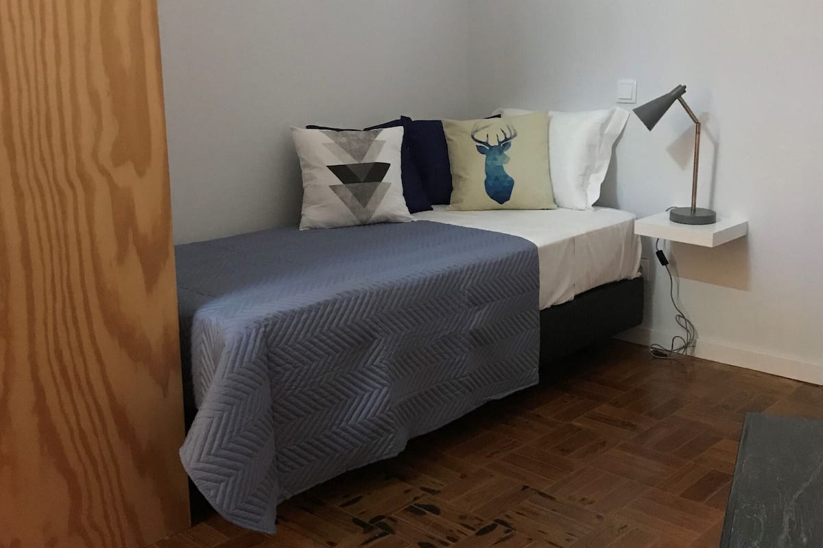 A small, neatly arranged bedroom with a bed, pillows, a bedside table, and a lamp.