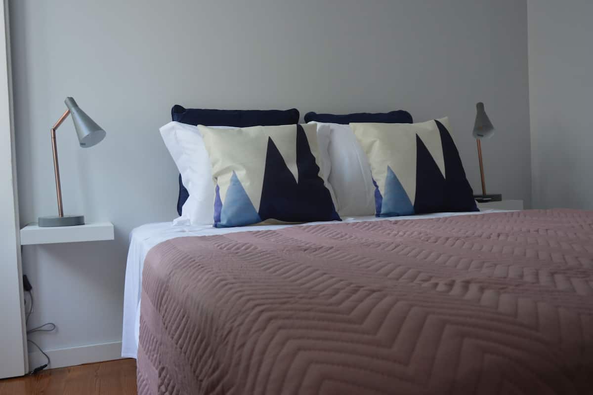 A neatly made bed with a pink quilt, two pillows with a mountain pattern, and two bedside lamps on white nightstands.