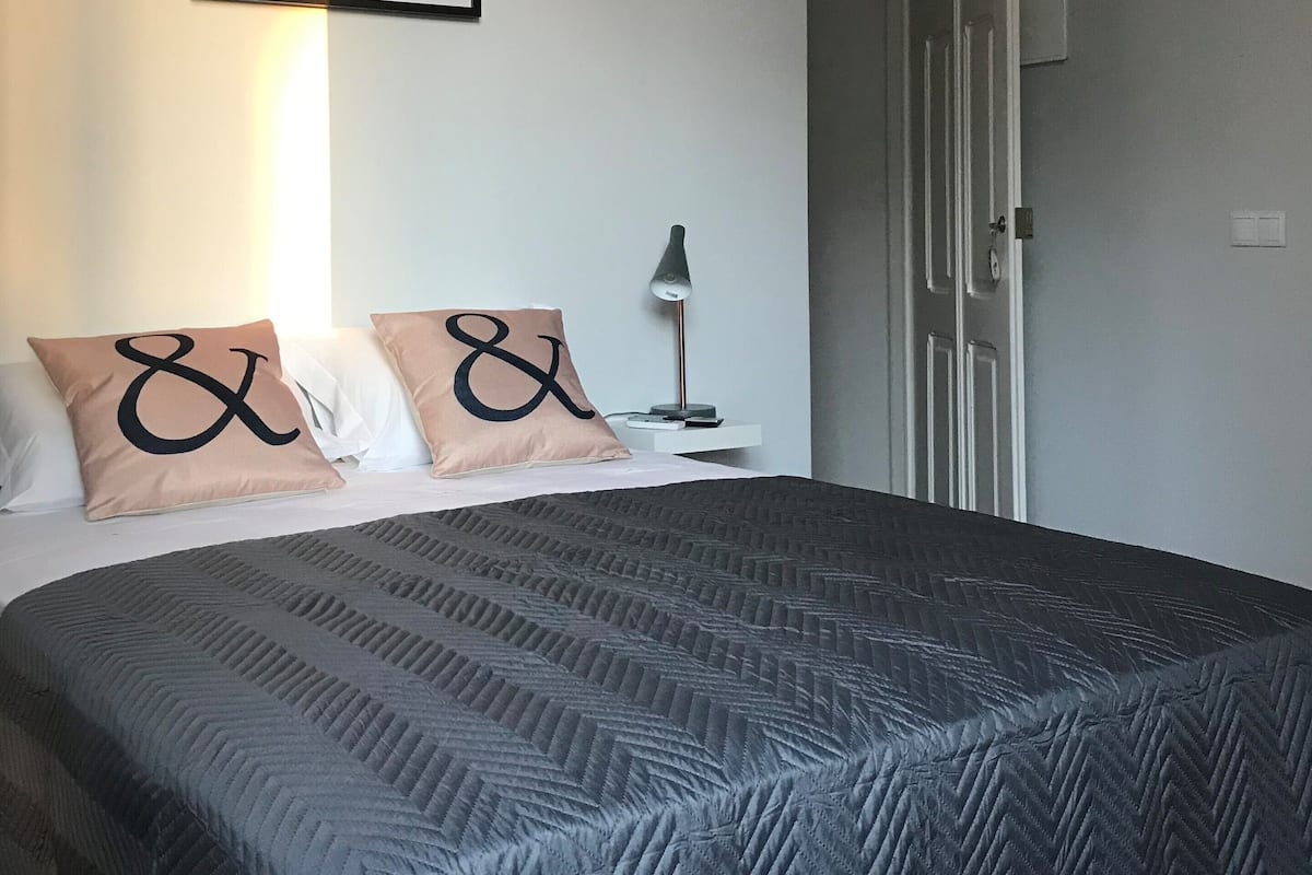 A neatly made bed with a patterned bedspread, two pillows with a large ampersand symbol, a framed artwork above the bed, a bedside lamp, and a door to the right.