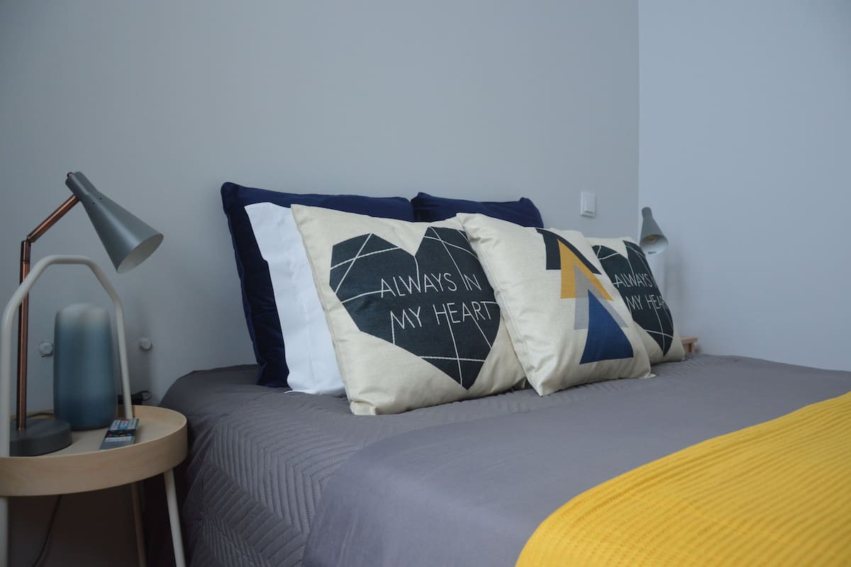 A neatly made bed with a grey and yellow blanket, two bedside lamps, and decorative pillows with a geometric design and text.