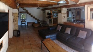 Un saló rústic amb un sofà de cuir negre, bigues de fusta, una televisió i una escala que condueix a una cuina.