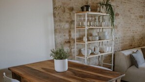 A rustic dining area with a wooden table and white chairs, a white shelving unit against a stone wall, and a sofa in the background. - La casa in piazzetta in Borgo Loretello (Loretello)