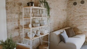A rustic room with exposed stone walls, a wooden ceiling, a white sofa with cushions, and a white shelving unit with glassware and plants. - La casa in piazzetta in Borgo Loretello (Loretello)