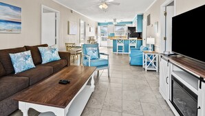 Un salón con un sofá marrón, sillones azules, una mesa de centro de madera y un televisor.