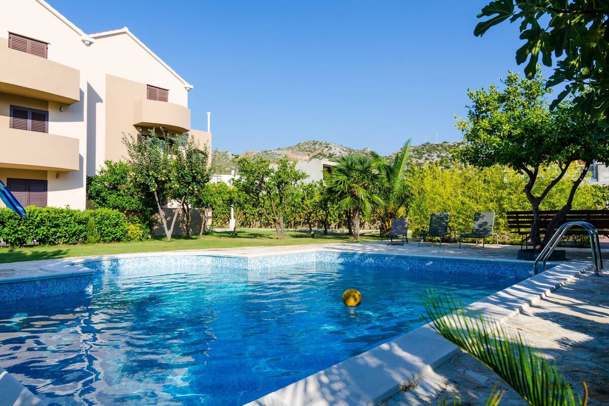 Une piscine à l’eau d’un bleu limpide, entourée de verdure et bordée d’un bâtiment doté de balcons.