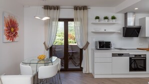 A modern kitchen with white cabinets, a built-in oven, and a microwave. There is a dining area with a glass table and white chairs. - Apartments Setemana (Zupa dubrovacka)