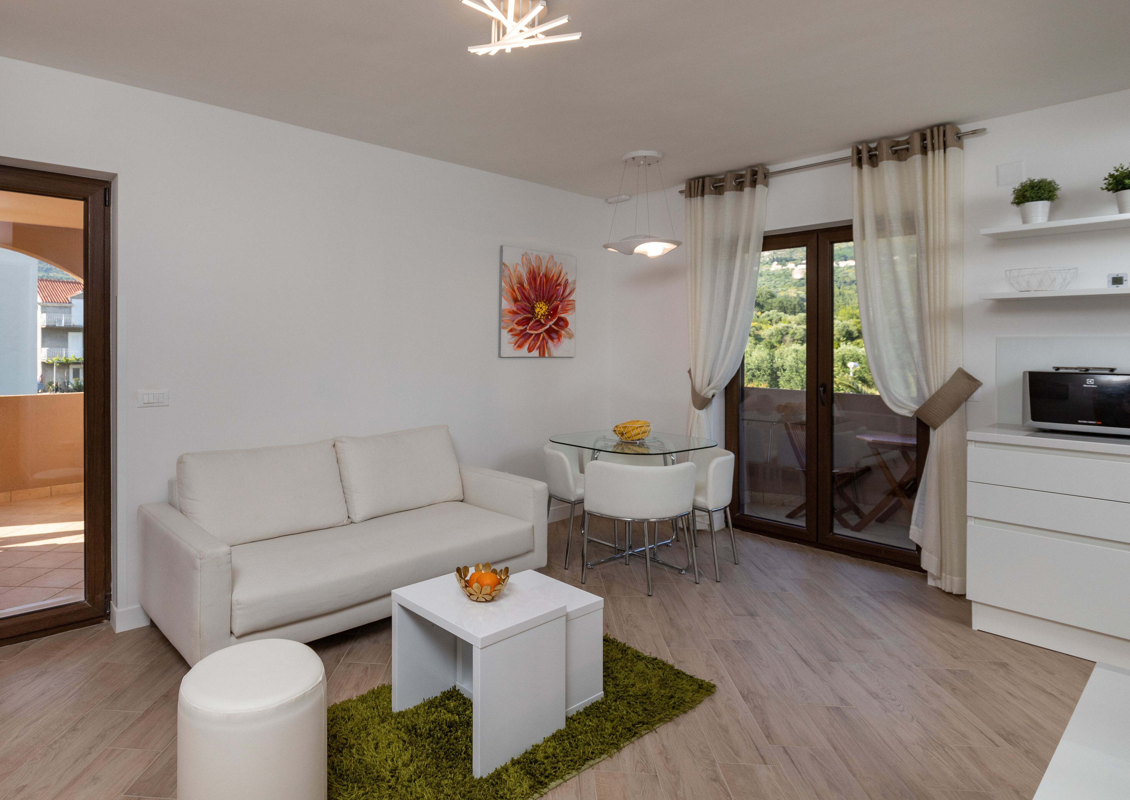 Un soggiorno moderno con un divano bianco, un tavolino in vetro, un tavolo da pranzo con sedie, una TV su un mobile e un balcone con vista.