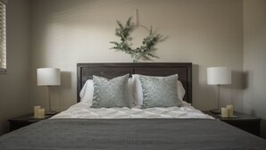 Un lit bien fait, recouvert d’une couverture grise, avec deux lampes de chevet et une guirlande décorative accrochée au mur.