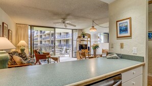 A modern kitchen with a large island, dining area with chairs and a table, and a balcony with a view of the building's interior. - Timeless island charm and breathtaking views (Lahaina)