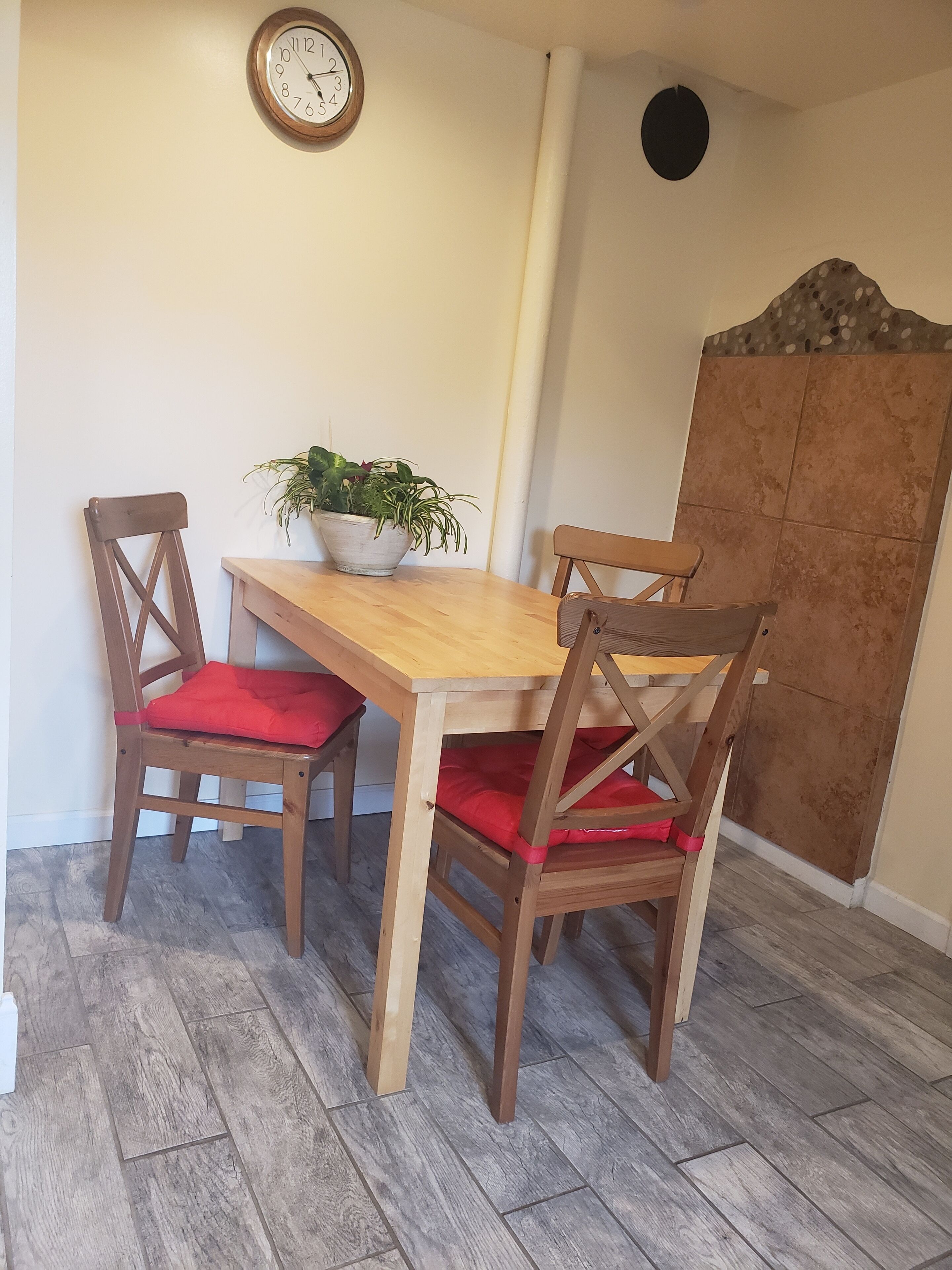 Een eetruimte met een houten tafel, vier stoelen met rode kussens, een potplant en een wandklok.