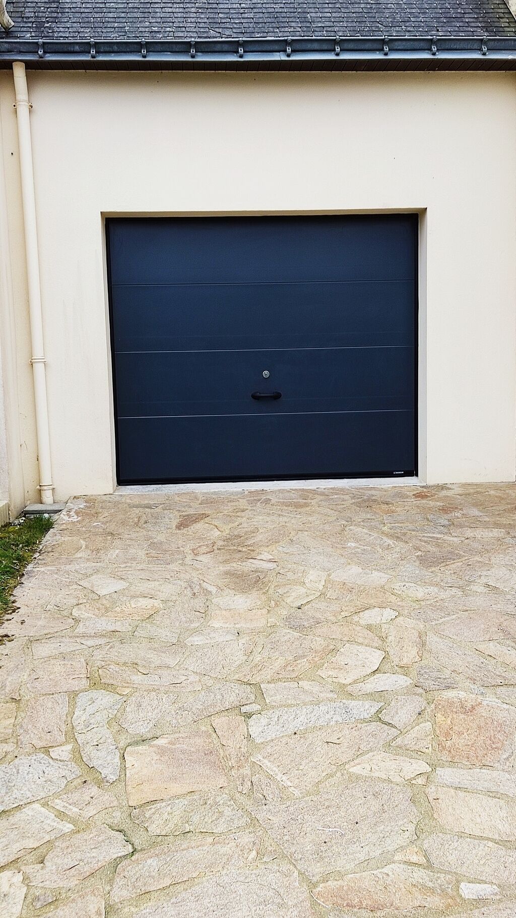 Un garatge amb una porta blava fosca, un camí d'accés pavimentat de pedra i una paret de color clara.