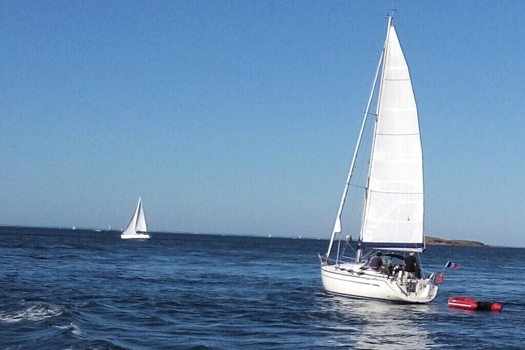 A sailboat on calm waters with a distant sailboat and a small red buoy.