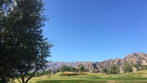 A golf course with rolling hills, a large tree, and mountains in the background.