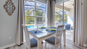 A dining area with a glass table, white chairs, and a view of a swimming pool through large windows. - POOL HOME - Luxury Windsor Palms Resort - 6 miles to Disney! POOL HEAT INCLUDED! (Kissimmee)