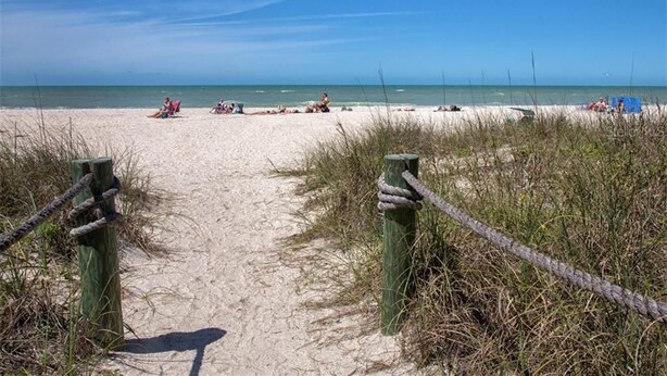Ein sandiger Küstenweg führt zum Meer, wo Menschen entspannen und Liegestühle stehen.