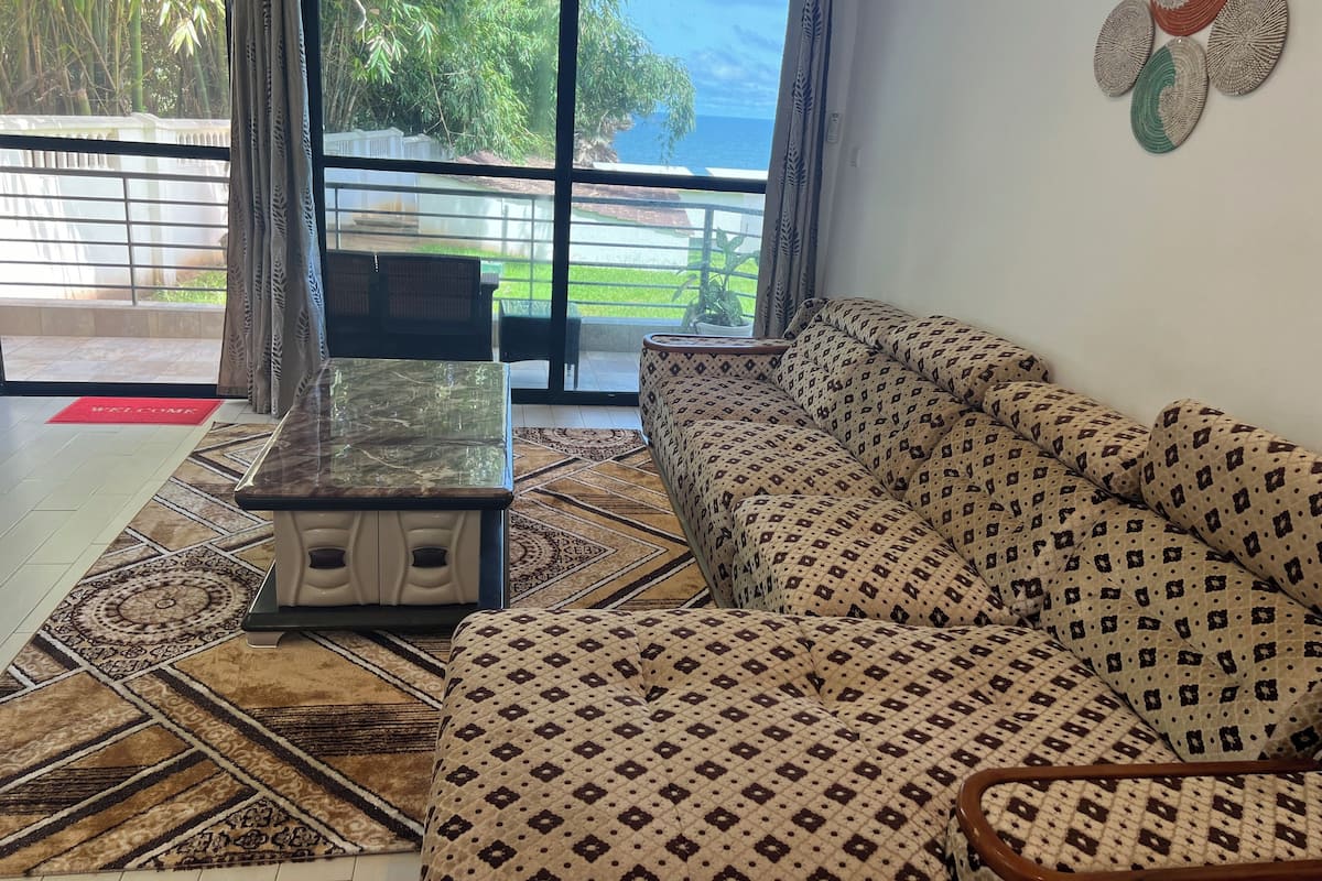 A living room with a patterned sofa, a coffee table, and a view of a balcony with a railing and greenery.