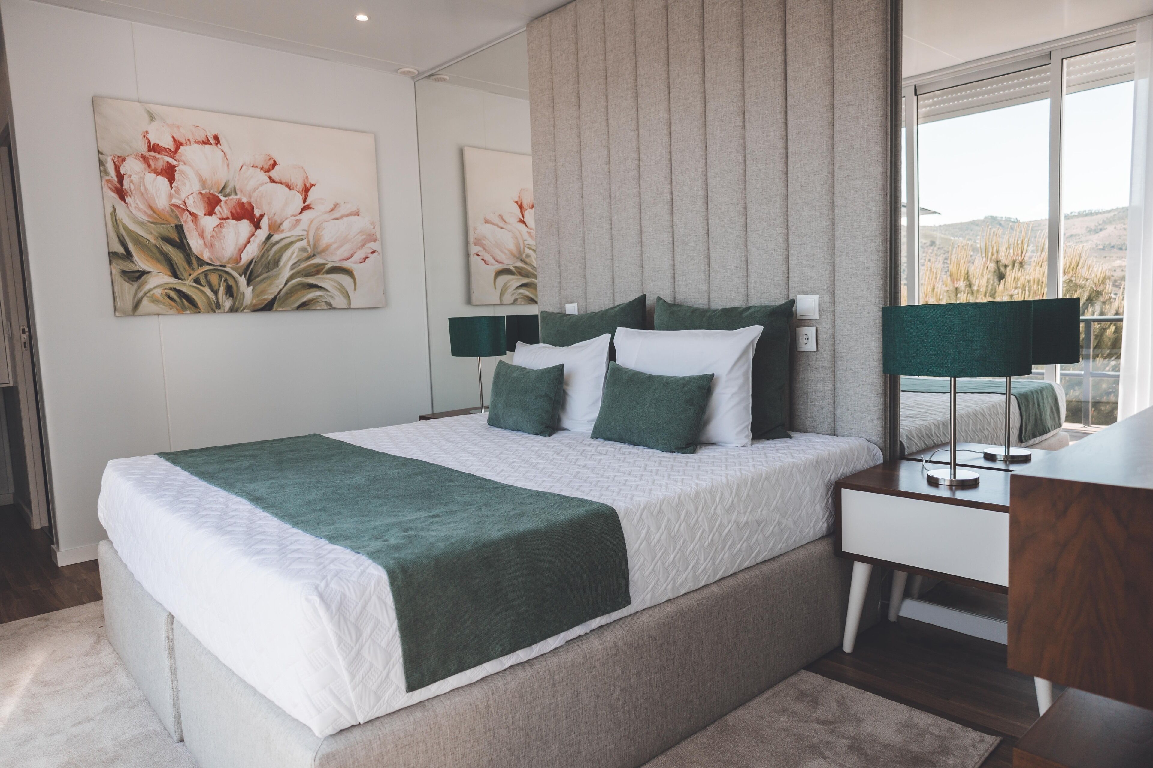 A modern bedroom with a large bed, green bedding, a bedside table, and a view of the outdoors.