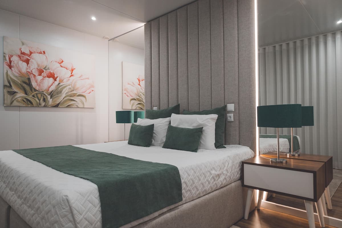 A modern hotel room with a large bed, green bedding, a nightstand with a lamp, and a wall-mounted painting.