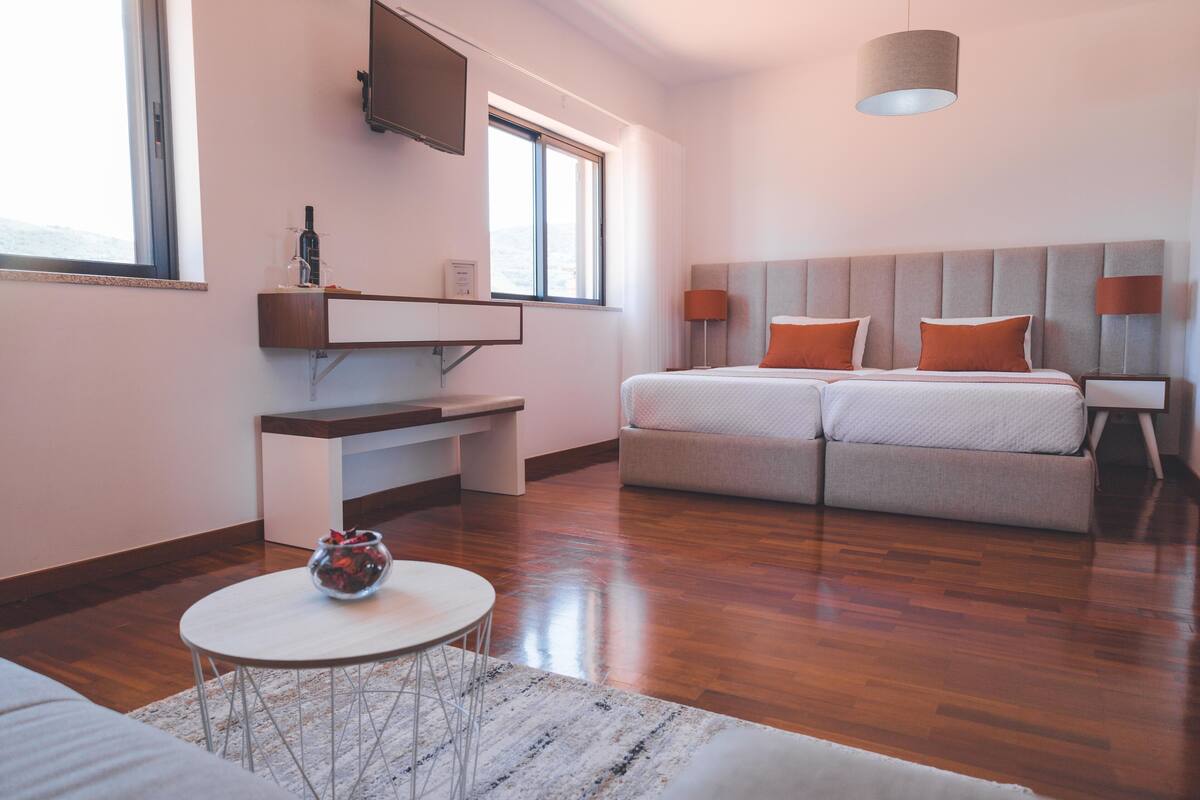 A modern hotel room with a sofa bed, a small table with a vase, a TV mounted on the wall, and a window providing natural light.