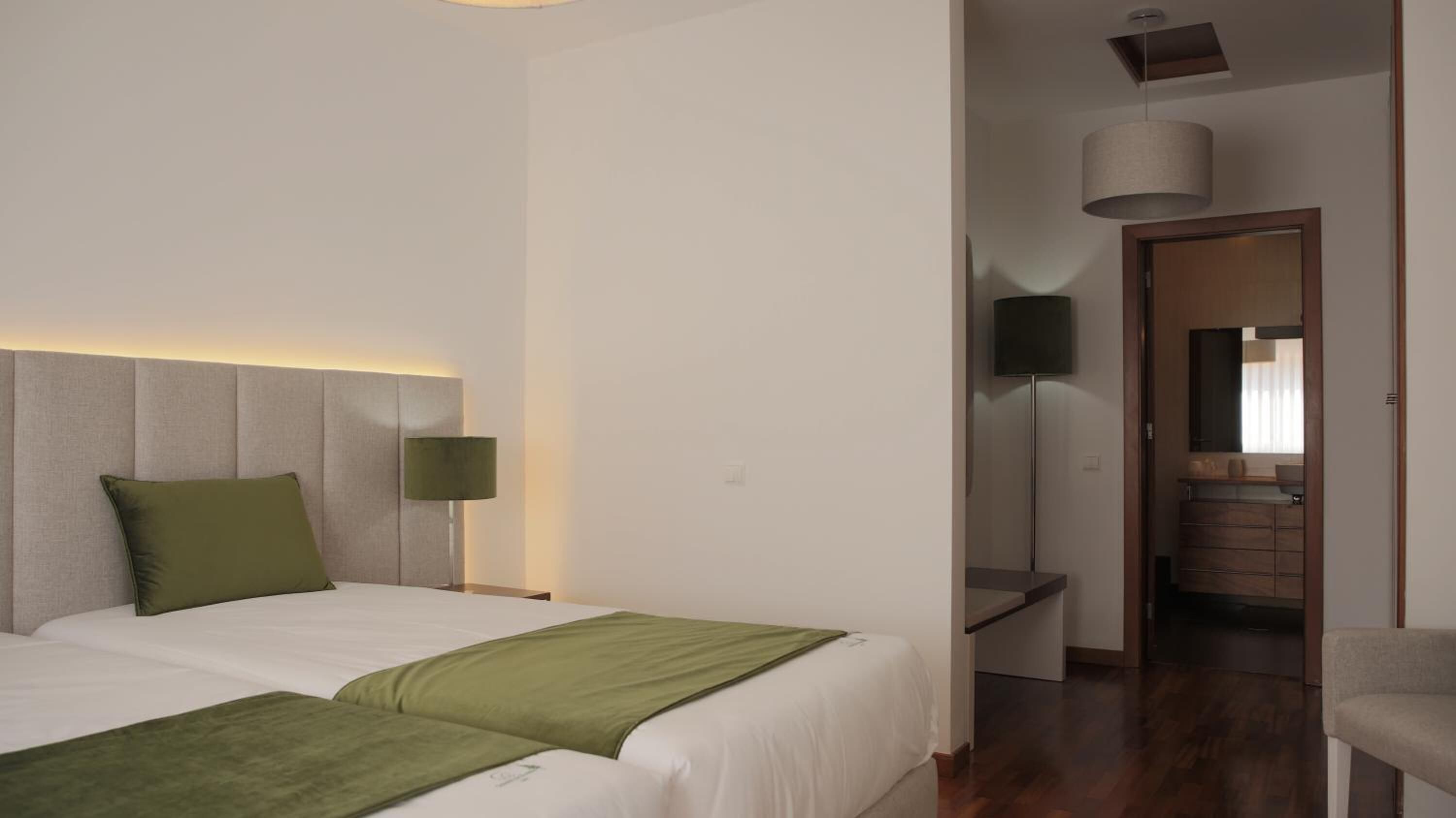 Ein Hotelzimmer mit zwei Betten, einem Kopfteil mit integrierter Beleuchtung, einem Nachttisch mit Lampe und Blick ins Badezimmer.